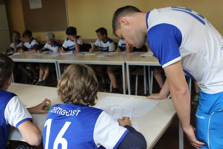 Kinder sitzend in einem Klassenraum, ein Muttersprachler hilft einem Kind bei den Aufgaben.